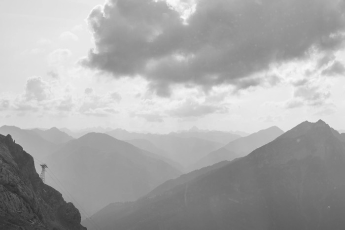Receding mountain ridges in haze with a lone cable-car pylon on a cliff and heavy cloud above