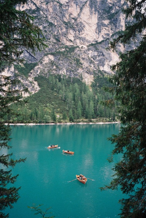 Three wooden rowing boats on a turquoise mountain lake framed by pine branches and cliffs