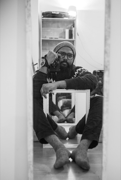 Bearded man in a beanie sits cross-legged on the floor with a camera to his eye, framed by a doorway and a small mirror