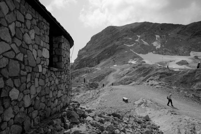 Stone-walled mountain hut at the left edge, hikers crossing the bare rocky slope beyond it