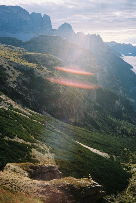 Steep alpine slope falling into a valley below jagged peaks, with a red light leak across the frame