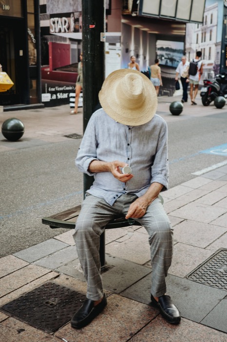 Man dozing on a street bench, straw hat tipped over his face, a coffee cup in his hand