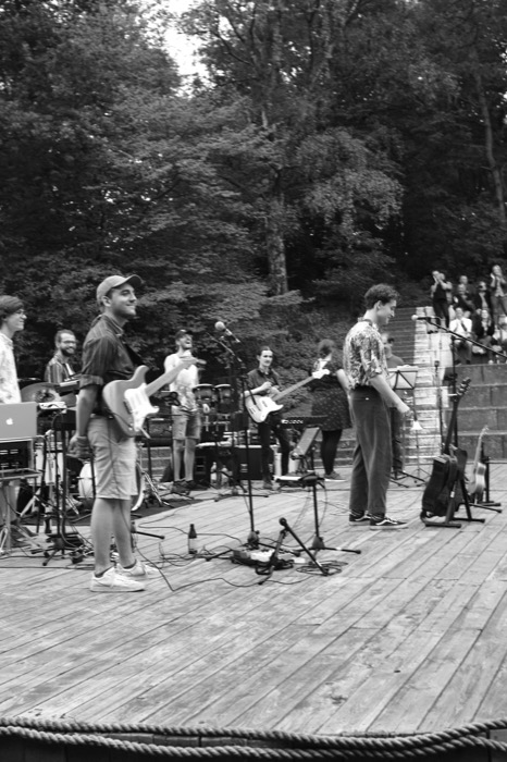 Band performing on an open-air wooden stage, guitarist mid-song, small crowd on the stone terraces