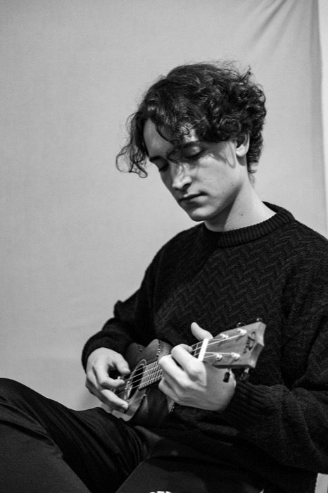 Curly-haired young man in a heavy knit sweater playing a ukulele, head bowed