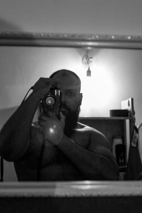 Shirtless bearded man photographing himself in a mirror, one eye visible past the camera, bulb flaring behind him