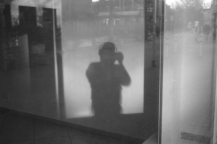Shadowy figure raising a camera, mirrored in a shop window with the empty street reflected beside