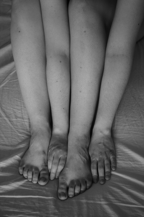 Bare legs and feet on a creased bedsheet with a hand laid across them, fingers mirroring the toes