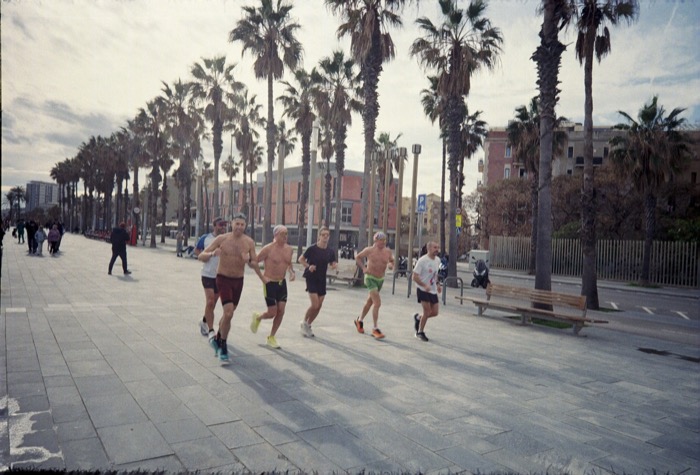 Group of shirtless runners jogging along a palm-lined seafront promenade