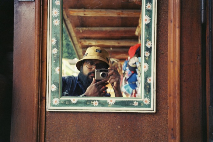 Bearded man in a bucket hat photographing himself in a daisy-painted mirror set in a wooden chalet door