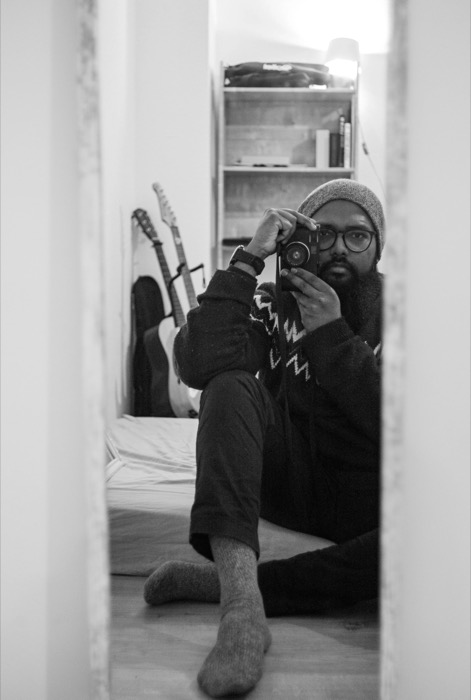 Bearded man in a beanie sits on a bedroom floor holding a camera to his eye, seen through a doorway
