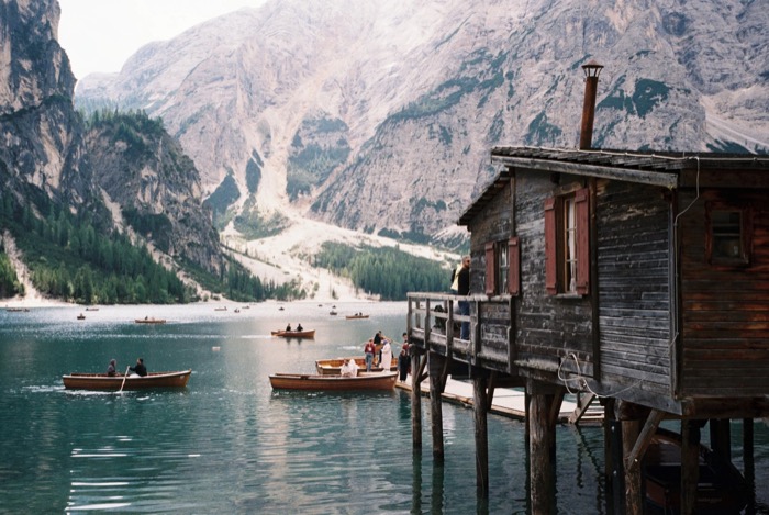 Wooden rowboats on a green mountain lake beside a weathered boathouse on stilts, cliffs behind