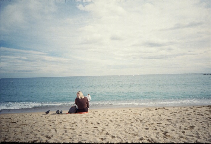 Lone woman sitting on a towel on an empty beach facing a calm sea under broken cloud