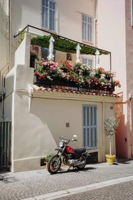 Red motorbike parked under a flower-filled balcony on a narrow sunlit street