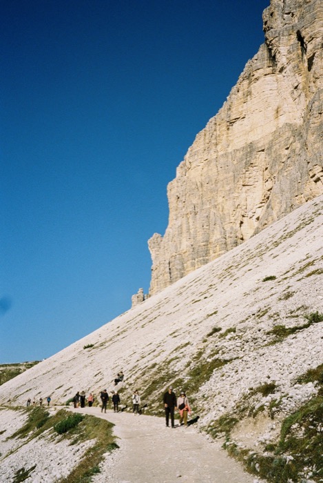 Hikers strung along a pale gravel path beneath a towering cliff face under deep blue sky