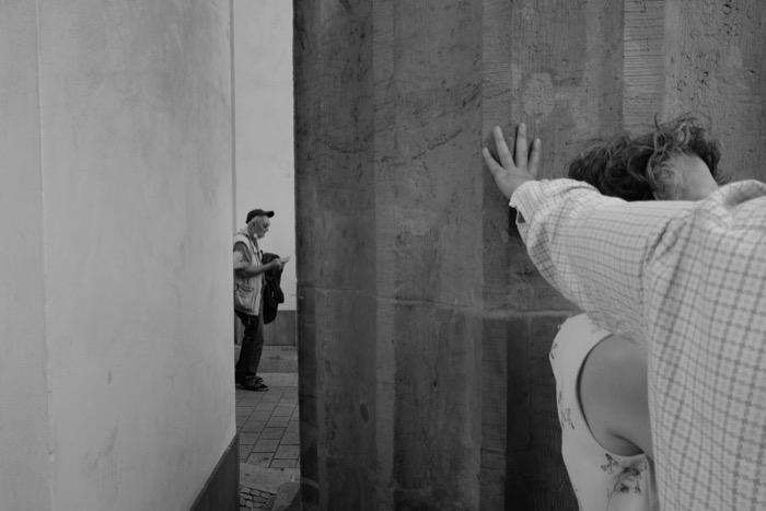 Woman pressing her palm and face to a stone column while a man in a cap reads in the alley beyond