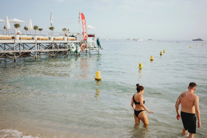 Two swimmers wading into calm sea beside a beach-club pier, yachts on the horizon