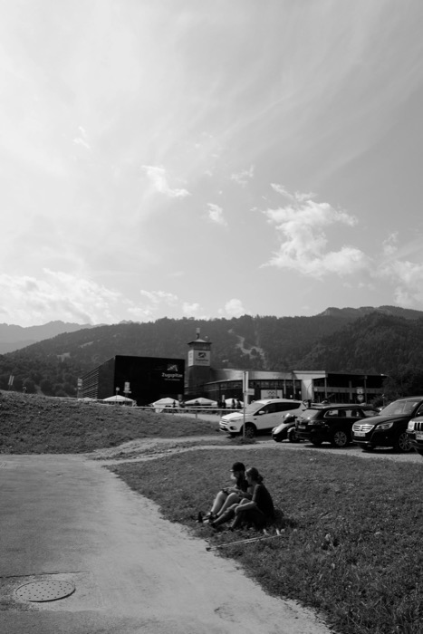 Two people sitting on a grass verge above a car park, wooded mountains and a cable-car station behind