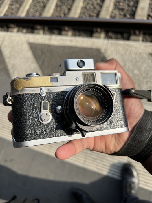 Hand holding a battered chrome rangefinder camera above a railway platform edge and tracks