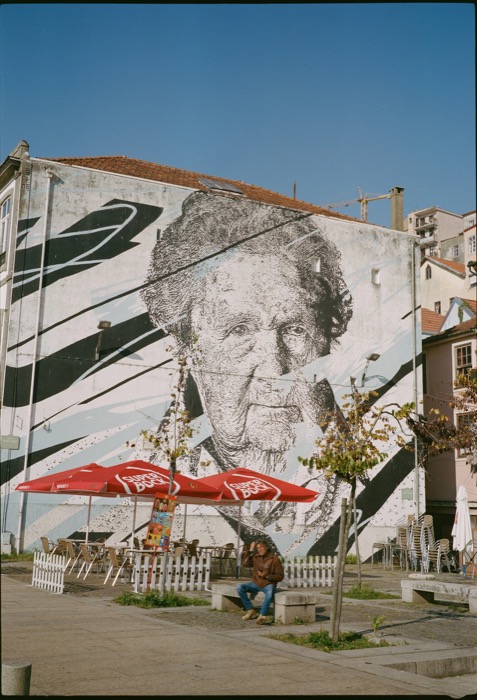 Huge mural of an elderly face on a gable wall above a cafe terrace with red parasols