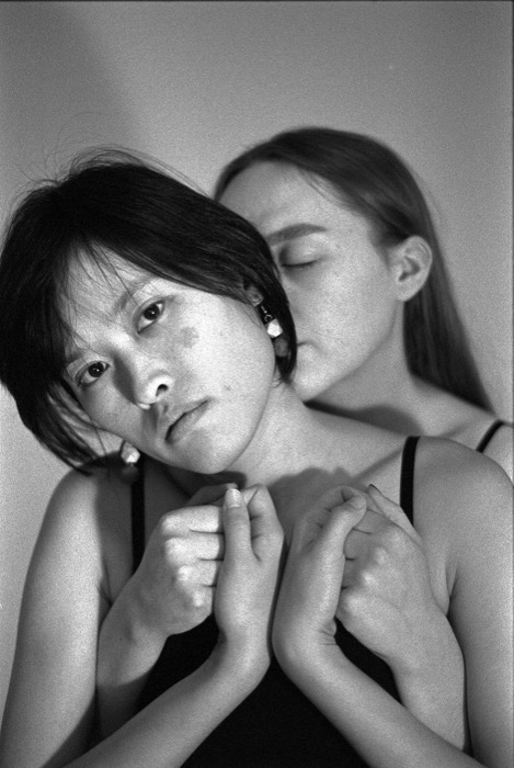 Two young women pressed close, one facing the lens with clenched hands at her chest, the other resting behind with eyes closed