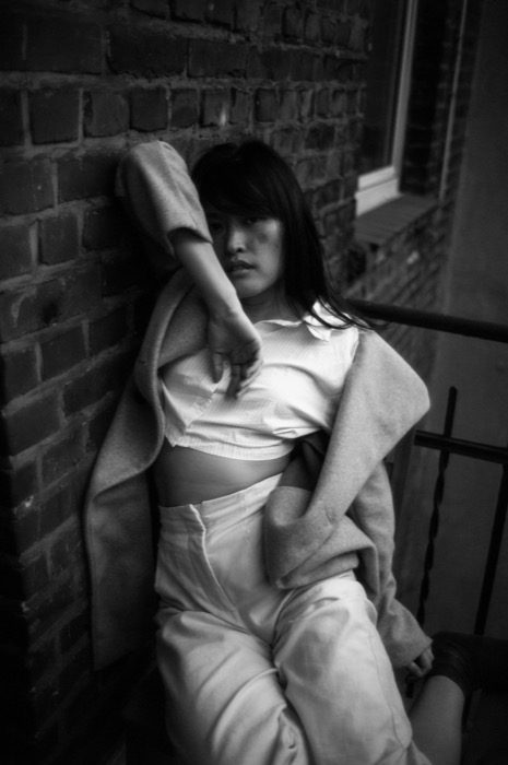 Woman in a coat and cropped shirt leaning back against a brick wall on a fire escape, arm raised overhead