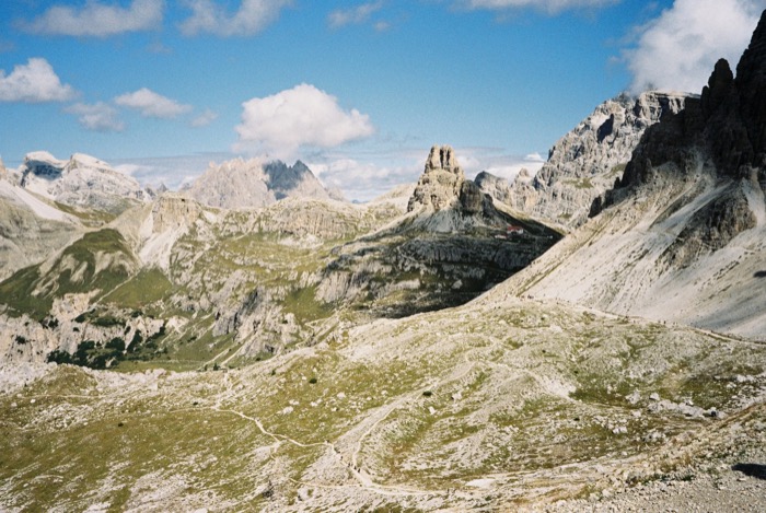 Wide mountain basin of pale scree and grass with a red-roofed hut and hikers on winding paths