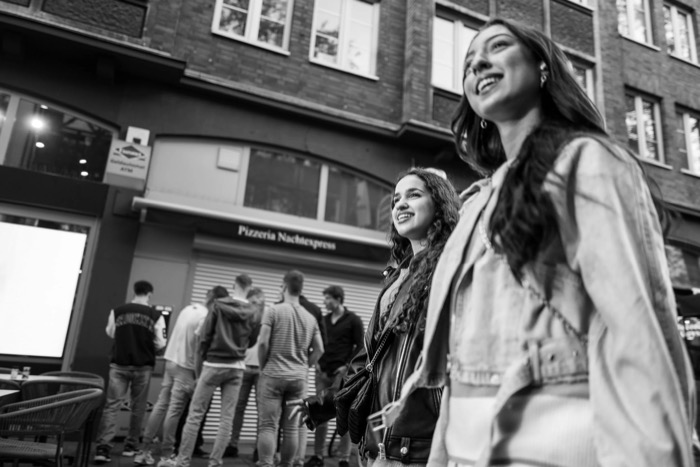 Two young women walking and laughing past a shuttered pizzeria, shot from below, group of men behind