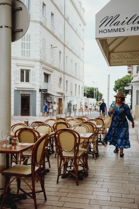 Woman in a blue floral dress and straw hat passing rows of empty rattan café chairs on a seaside street