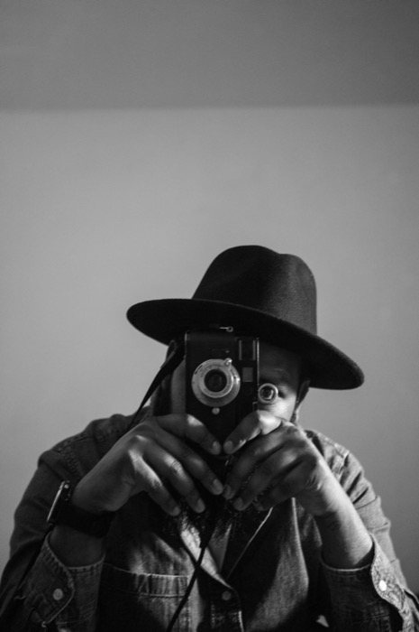 Self-portrait in a wide-brimmed hat with a rangefinder camera hiding the face against a plain wall