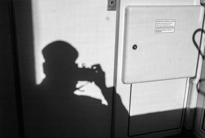 Shadow of a person in a cap raising a camera, cast on a train wall beside a handbrake box