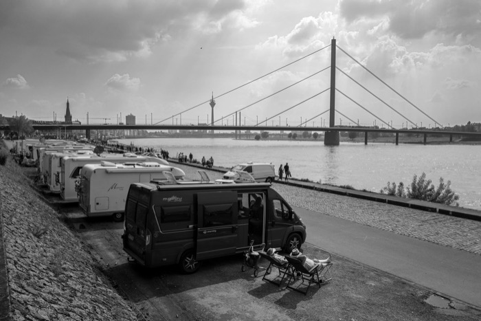 Row of campervans parked along a riverbank promenade, a cable-stayed bridge and TV tower beyond