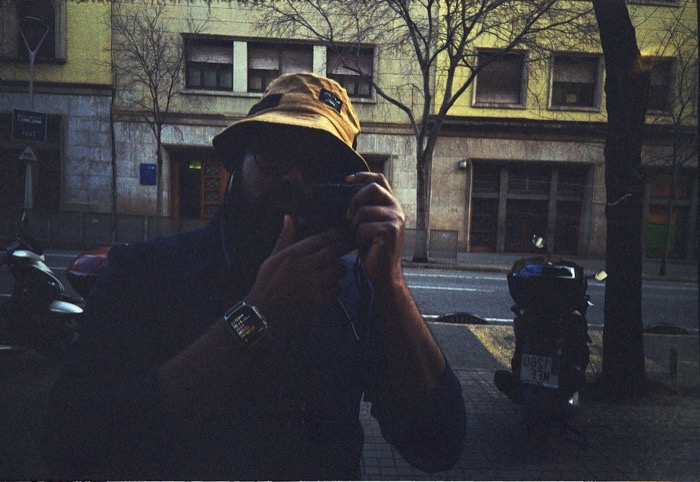 Reflection in a dark window of a bearded man in a yellow bucket hat raising a compact camera, street behind