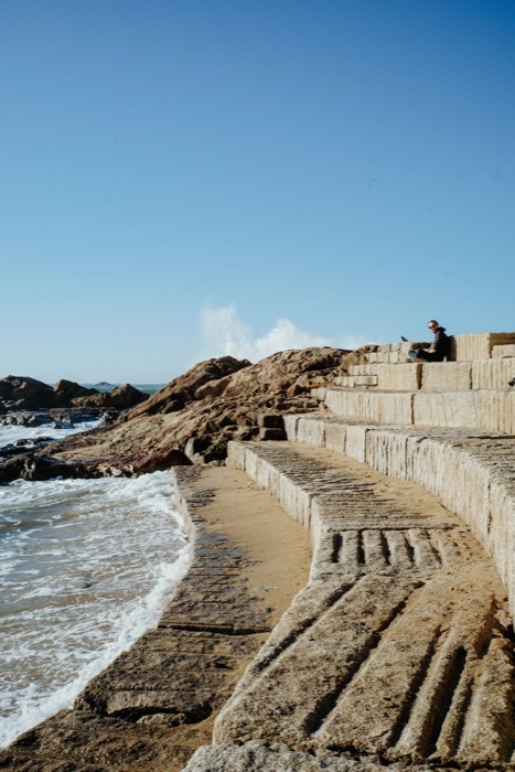Figure sitting alone on a stepped stone breakwater above surf, under a wide empty blue sky