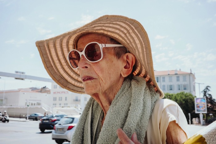Elderly woman in a wide straw sun hat, white sunglasses and a towel round her neck on a sunlit street