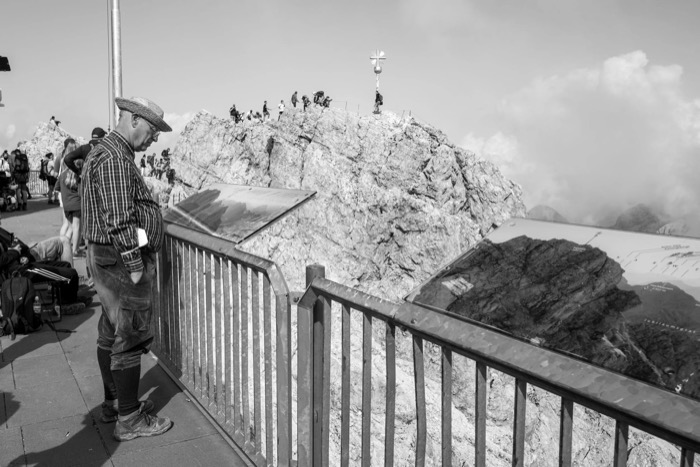 Man in checked shirt and knee socks at a railing, looking toward tourists crowding a summit cross on bare rock
