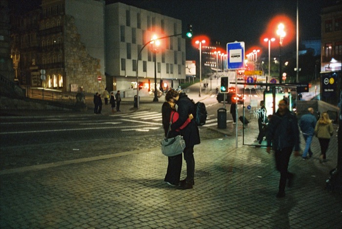 Couple embracing on an empty cobbled square at night, city lights and passers-by around them