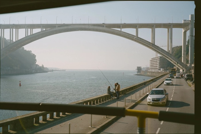 Anglers on a riverside wall beneath a vast concrete arch bridge, traffic passing on the road alongside