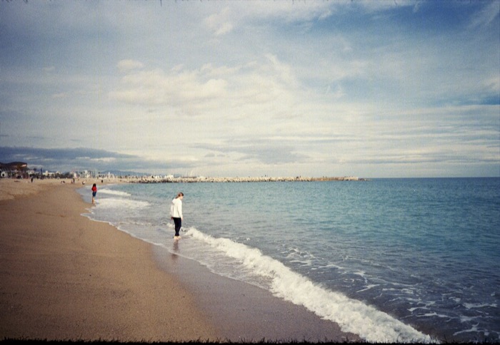 Girl standing barefoot in the shallow surf on a wide empty beach under a big cloud-streaked sky