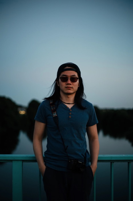 Long-haired man in sunglasses and a backwards cap standing on a bridge railing at dusk, camera slung at his hip