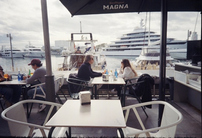 Couple talking at a marina cafe table under a parasol, yachts and a large white ship behind