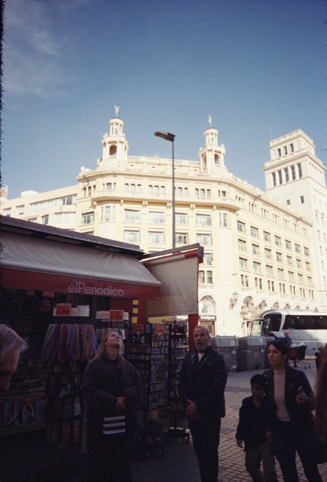 Passers-by at a newspaper kiosk marked el Periodico, one man staring up at the grand building above