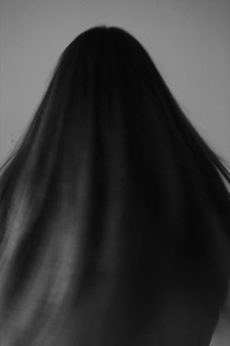 Back of a head of long dark hair filling the frame against a pale wall
