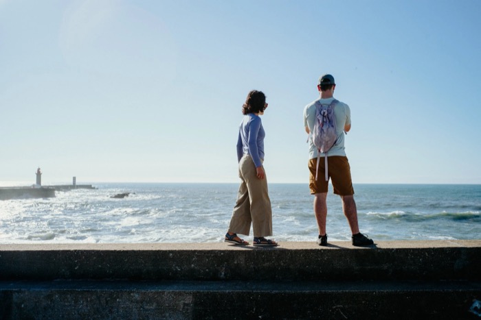 Couple standing on a sea wall looking out at breaking waves, a lighthouse on the jetty to the left