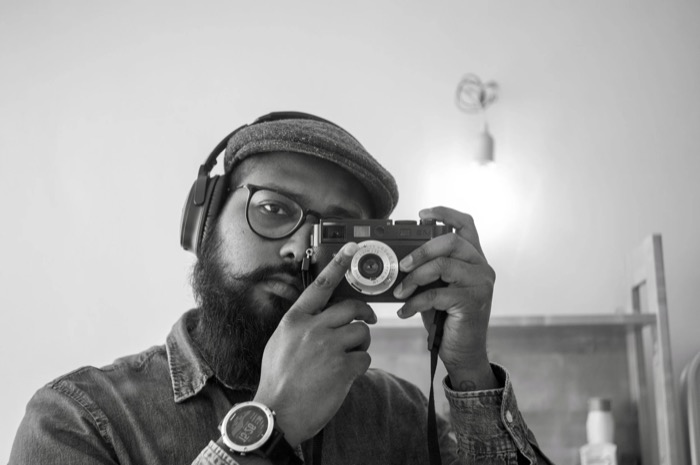 Bearded man in flat cap, glasses and headphones raises a rangefinder camera to his eye in a bare room
