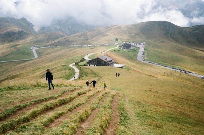 Walkers on a grassy alpine ridge above a mountain lodge and a long line of parked cars