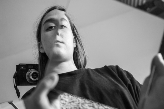 Low-angle self-portrait of a long-haired person reaching toward the lens, a film camera on the wall behind
