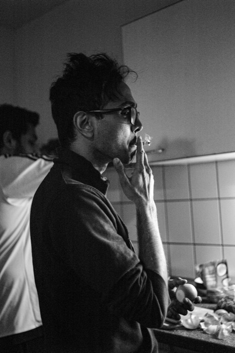 Man in glasses smoking in profile in a tiled kitchen, another person just behind him holding an onion