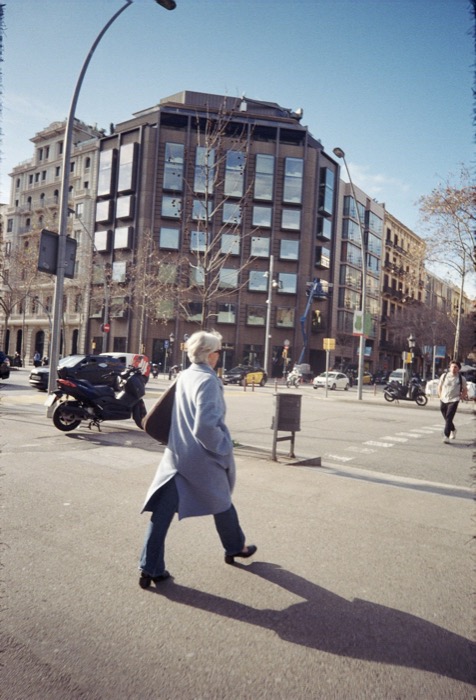 White-haired woman in a pale coat striding across a sunlit avenue, long shadow trailing behind her