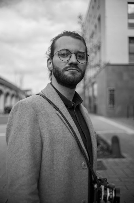 Bearded man in round glasses and a pale wool coat stands on an empty street, camera on a strap