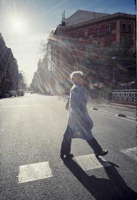 White-haired woman in a long coat striding across an empty crosswalk into strong backlight, sun flaring